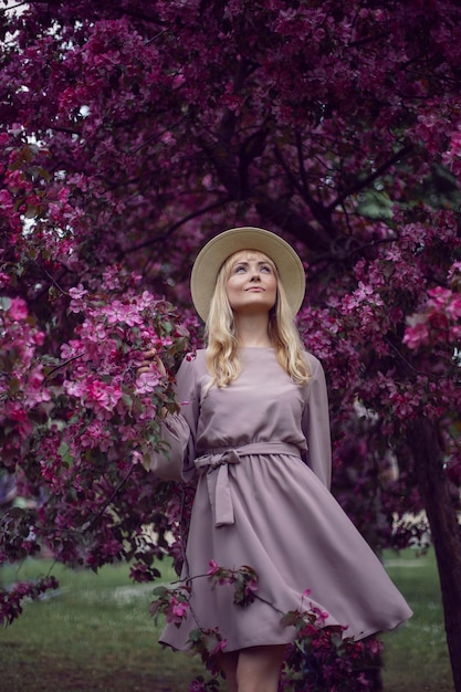 Portret jonge mooie vrouw in een roze jurk en hoed staat bij een bloeiende roze appelboom