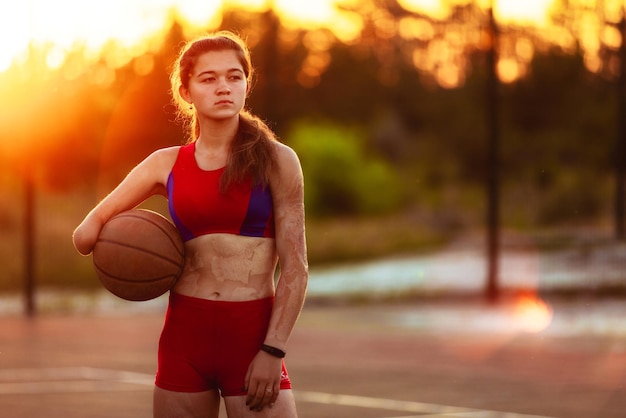 Portret jonge vrouw atleet met een geamputeerde arm en brandwonden op haar lichaam ze houdt een basketbalbal vast na een training buiten bij zonsondergang