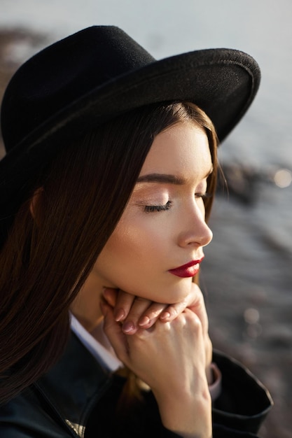 Portret jonge vrouw bij zonsondergang. Mode vrouw in zwarte ronde hoed en leren regenjas in de buurt van de vijver van het meer. Mooie make-up en felrode lippenstift op de lippen