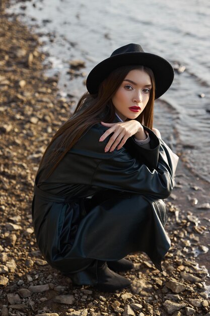 Portret jonge vrouw bij zonsondergang. Mode vrouw in zwarte ronde hoed en leren regenjas in de buurt van de vijver van het meer. Mooie make-up en felrode lippenstift op de lippen