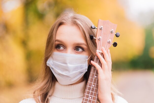 Portret jonge vrouw in beschermend masker ukelele gitaar spelen in herfst park, levensstijl gezond reizen concept