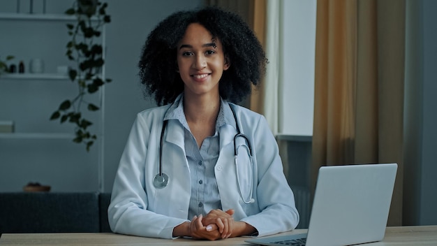 Portret lachende Afrikaanse dokter vrouw dermatoloog in medisch uniform zit in kliniek