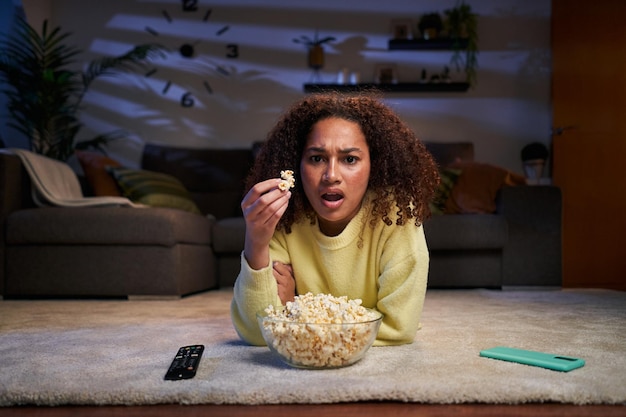 Portret latina verrast afro vrouw liggend op woonkamer tapijt eten popcorn uit de schaal geschokt