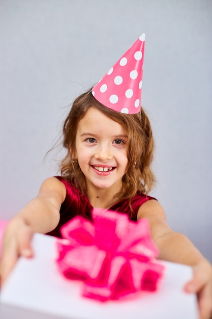 Portret leuk glimlachend meisje die in verjaardagshoed een roze en witte giftdoos houden