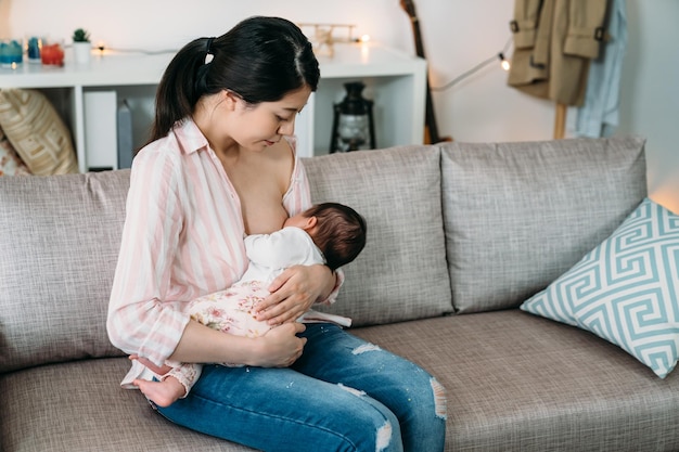 portret liefhebbende aziatische moeder kijkt en geeft haar babymeisje borstvoeding in haar wieg op de bank in de woonkamer thuis