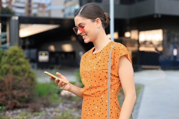 Portret mode vrouw in gele jurk in zonnebril lopen op straat en smartphone in haar hand houden.