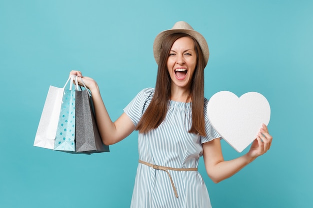 Portret modieuze mooie vrouw in zomerjurk, strohoed met pakketten tassen met aankopen na het winkelen, wit hart met kopie ruimte voor reclame geïsoleerd op blauwe pastel achtergrond.