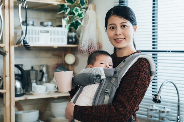 portret mooie aziatische moeder die thee vasthoudt, lacht naar de camera terwijl ze een onschuldige baby draagt met een draagdoek in de ochtend op een moderne, lichte keukenachtergrond