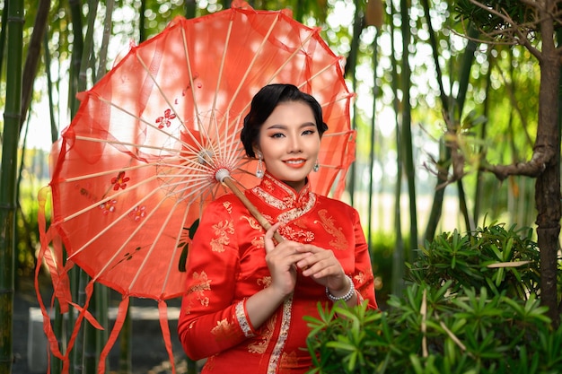 Portret mooie Aziatische vrouw in een Chinese cheongsam poseren met mooie rode papieren paraplu, glimlach en op zoek naar camera op bamboebos, kopieer ruimte