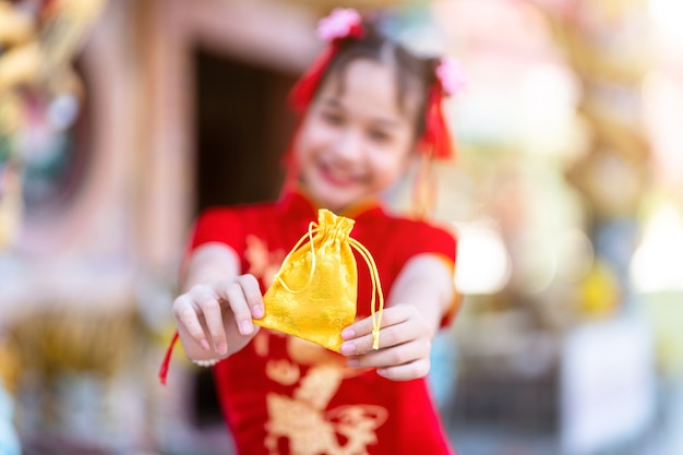 Portret mooie glimlach schattig klein aziatisch meisje dragen rode traditionele chinese cheongsam, focus toon gouden geld tas voor chinees nieuwjaar festival op chinees heiligdom