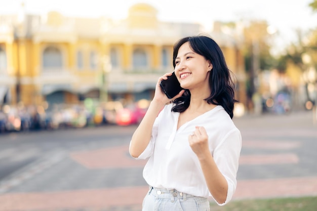 Portret mooie jonge aziatische vrouw die goed nieuws ontvangt op mobiele telefoon en opgeheven armen voelt vrolijk geluk rond buiten straatbeeld in een zomerse dag