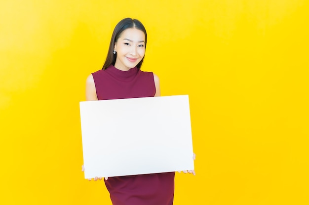 Portret mooie jonge aziatische vrouw met leeg wit reclamebord op gele muur