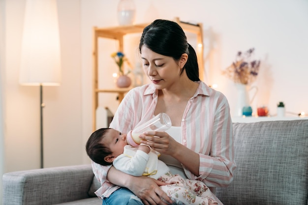 portret mooie liefdevolle aziatische nieuwe moeder wiegt en voedt haar baby overdag met flesmelk op de bank in een gezellige woonkamer thuis.