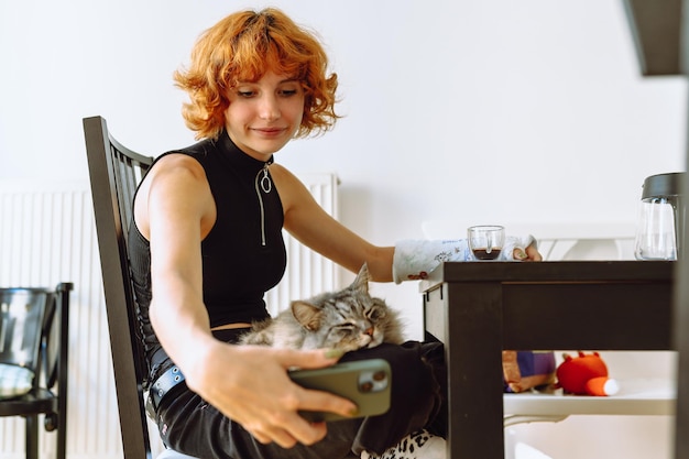 Portret roodharig tienermeisje met kat in haar armen over een kop koffie met een telefoon thuis