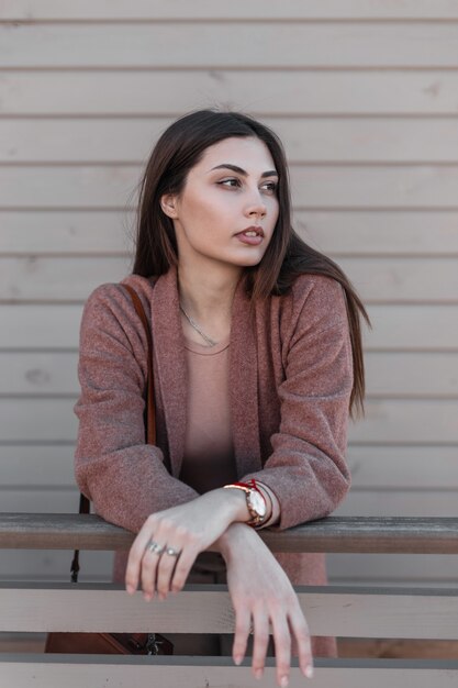 Portret sensuele aantrekkelijke bruinharige jonge vrouw in modieuze vintage jas met sexy lippen in de buurt van houten gebouw in de stad. Aantrekkelijk meisje in mooie elegante kleding rust buiten op lentedag.