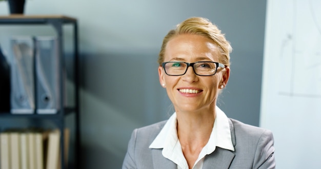 Foto portret shot van kaukasische glimlachte mooie vrouw in glazen glimlachend naar camera zittend in kantoor. close-up van knappe zakenvrouw in kabinet. vrouwelijke leraar op school.