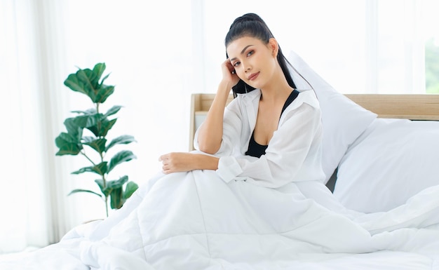 Portret shot van Millennial Aziatische sexy vrouw in casual comfort tanktop en shirt zittend onder een witte dikke warme deken poseren kijken camera glimlachen na het wakker worden in de slaapkamer in de ochtend thuis