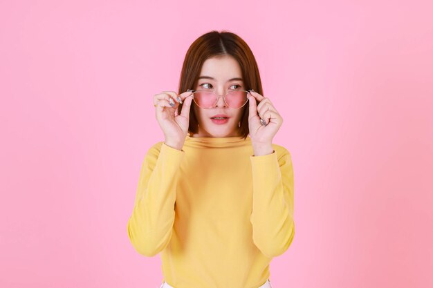 Portret studio knipsel shot Aziatische jonge mooie kort haar vrouwelijke stijlvolle modieuze model in gele shirt met lange mouwen staande kijken naar camera bedrijf mode zonnebril op roze achtergrond aan te passen.