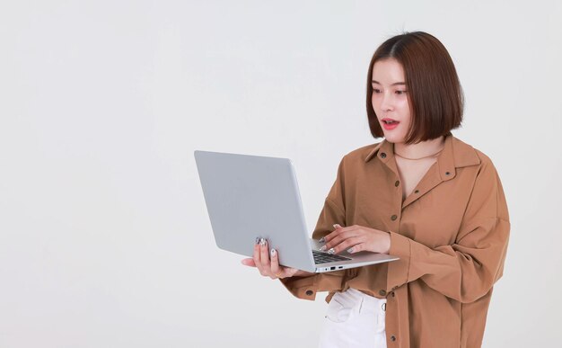 Portret studio knipsel shot Aziatische jonge mooie kort haar vrouwelijke student model in lang bruin shirt staande glimlachend bedrijf typen browsen surfen leren van laptopcomputer op witte achtergrond.