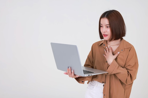 Portret studio knipsel shot Aziatische jonge mooie kort haar vrouwelijke student model in lang bruin shirt staande glimlachend bedrijf typen browsen surfen leren van laptopcomputer op witte achtergrond.