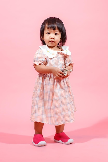 Portret studio shot kleine schattige Aziatische kleuterschool kleuter meisje dochter model in casual lange jurk en schoenen staande kijken naar camera met honderd dollar biljet in handen op roze achtergrond.