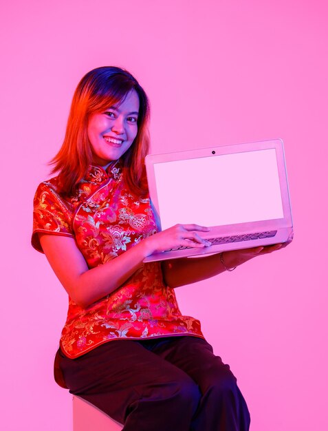 Portret studio shot millennial Aziatisch vrouwelijk model in het rood Chinese traditionele cheongsam qipao shirt zitten bedrijf tonen presenteren leeg scherm laptop computer product op witte achtergrond.