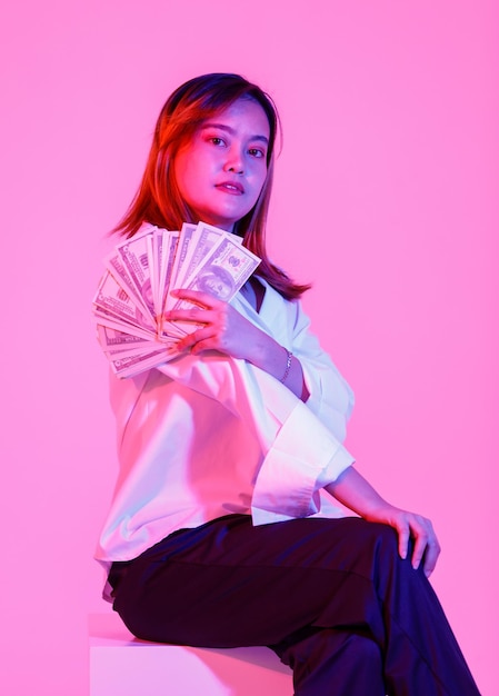 Portret studio shot van aziatische rijke rijkdom vrouwelijke zakenvrouw model in casual draagt zittend kijken naar camera met honderd dollar usd contant bankbiljet valuta stapel op roze achtergrond.