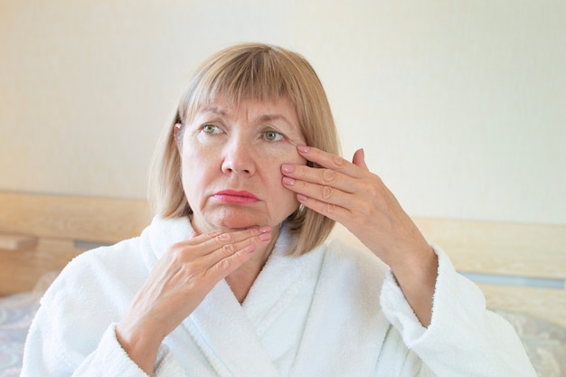 Portret triest bejaarde achtergrond van zijn slaapkamer. Haar handen raken de rimpels in haar gezicht. Concept Anti-leeftijd, gezondheidszorg en cosmetologie, vermoeidheid, ouderdom en ziekte denken