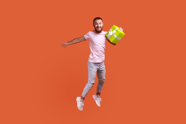 Portret van aantrekkelijke bebaarde man springen in de lucht met huidige doos in handen kijken camera kopie ruimte voor advertentie dragen roze tshirt en jeans Indoor studio shot geïsoleerd op oranje achtergrond