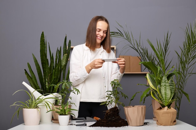 Portret van aantrekkelijke bruinharige blanke vrouw botanicus die in haar oranjerie werkt en foto's maakt op mobiele telefoon voor haar blog over thuisbroeken die voor bloemen zorgen