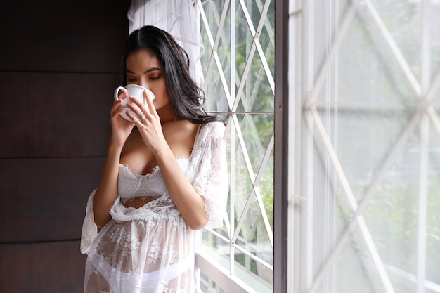 Portret van aantrekkelijke en sexy Aziatische vrouw in witte lingerienachtkleding die zich naast venster bevinden en koffie in de ochtend met ontspanning drinken.