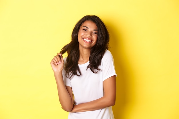 Portret van aantrekkelijke en zelfverzekerde Afro-Amerikaanse vrouw die een witte t-shirt draagt die gelukkig glimlacht en met haar speelt