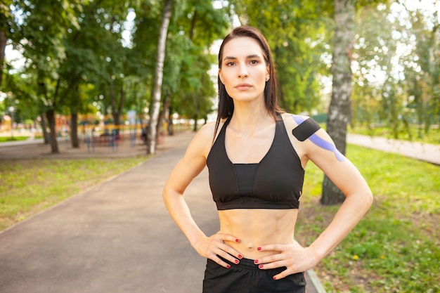 Portret van aantrekkelijke gespierde brunette vrouw, gekleed in zwarte sport outfit, camera kijken. jonge lachende vrouwelijke atleet poseren met handen op de taille, kleurrijke kinesiotaping op lichaam.