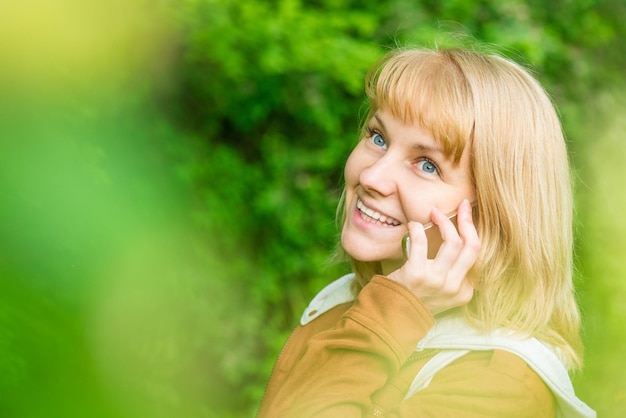 Portret van aantrekkelijke glimlachende blanke jonge vrouw met mobiele telefoon in het voorjaarspark die van dichtbij wegkijkt Mooi meisje dat aan het praten is op de mobiele telefoon