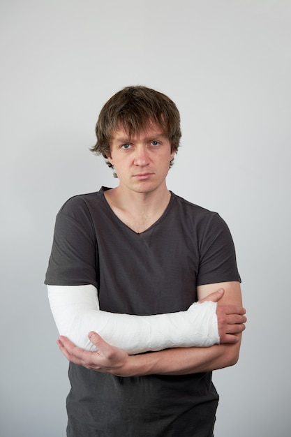 Foto portret van aantrekkelijke jonge boos blanke man met gipsverband op zijn hand op een witte muur achtergrond.