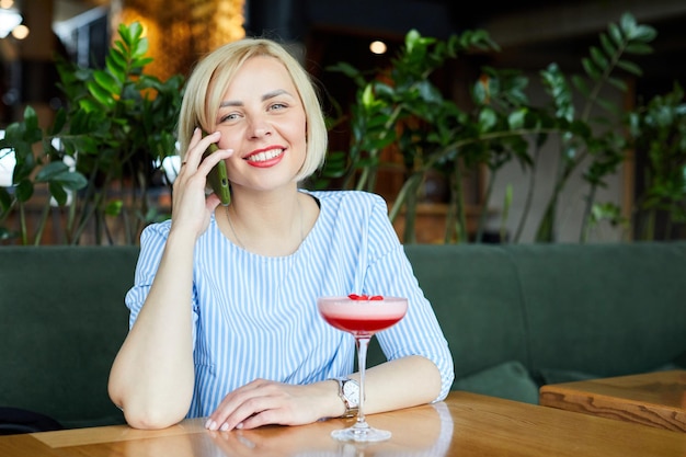 Portret van aantrekkelijke jonge vrouw cocktail drinken in café binnen Mooie blonde vrouw ontspannen aan de bar en een cocktail drinken en praten aan de telefoon