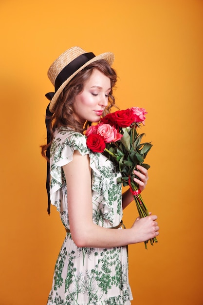 Portret van aantrekkelijke jonge vrouw in zomerjurk en strohoed met rozenboeket op gele achtergrond