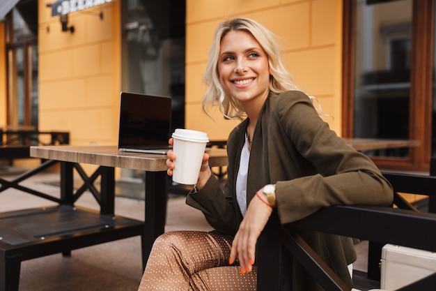 Portret van aantrekkelijke jonge vrouw met afhaalkoffie, en met behulp van zilveren laptop zittend in café buiten