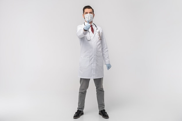 Portret van aantrekkelijke knappe dokter in beschermend gezichtsmasker, witte laboratoriumjas, stropdas geïsoleerd op een witte muur