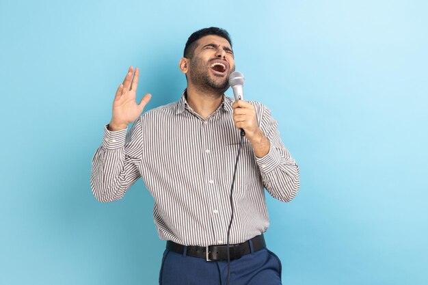 Portret van aantrekkelijke knappe jonge volwassen man met baard luid favoriete liedjes zingen met microfoon in handen dragen gestreept shirt Indoor studio opname geïsoleerd op blauwe achtergrond