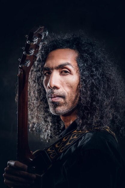 Portret van aantrekkelijke krullende man met gitaar in donkere fotostudio.