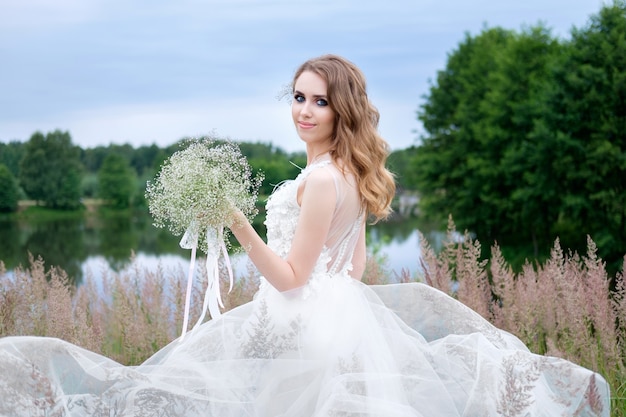 Portret van aantrekkelijke lachende jonge stijlvolle bruid in witte trouwjurk met bruiloft boeket in de hand, make-up en kapsel