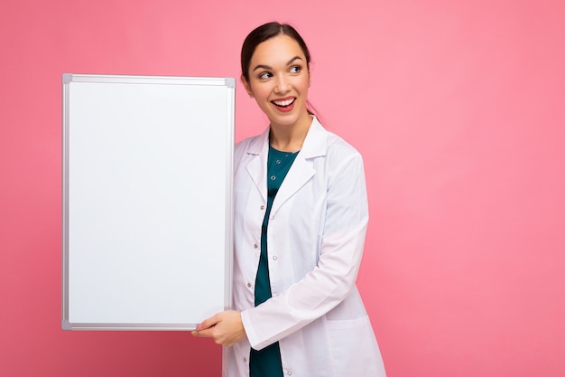 Portret van aantrekkelijke mooie jonge positieve brunette vrouw, gekleed in medische witte jas met wit magnetisch bord voor mock up geïsoleerd op roze achtergrond met kopie ruimte.