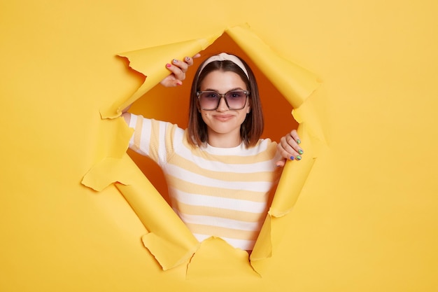 Portret van aantrekkelijke positieve vrouw die haarband en gestreept t-shirt draagt dat zich in geel document gat bevindt dat camera met brede glimlach bekijkt die in goed humeur is