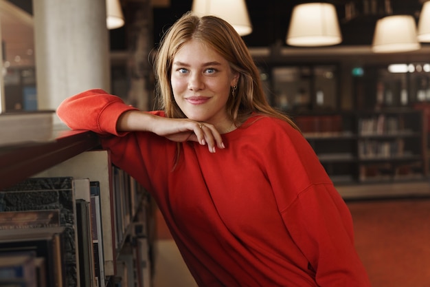 Portret van aantrekkelijke roodharige vrouwelijke student, leunend op een boekenplank in een bibliotheekzaal en glimlachende camera.