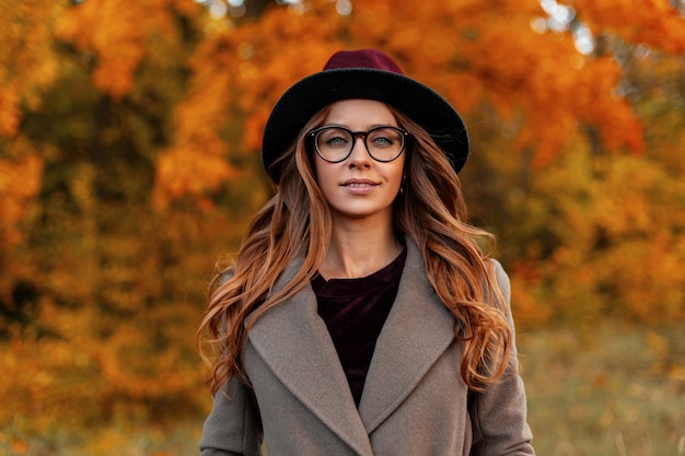 Portret van aantrekkelijke stijlvolle jonge vrouw in trendy bril in een vintage hoed in een elegante jas op een achtergrond van een bomen met gouden bladeren in het park. Mooi Europees hipster meisje buitenshuis.