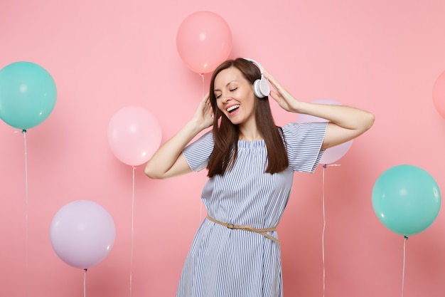 Portret van aantrekkelijke vrolijke jonge vrouw met gesloten ogen met koptelefoon in blauwe jurk luisteren muziek op roze achtergrond met kleurrijke luchtballonnen. Verjaardag vakantie feest mensen oprechte emoties.