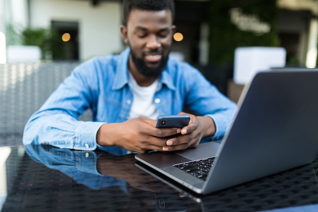 Foto portret van afrikaanse man praten aan de telefoon met laptop voor hem op tafel in café