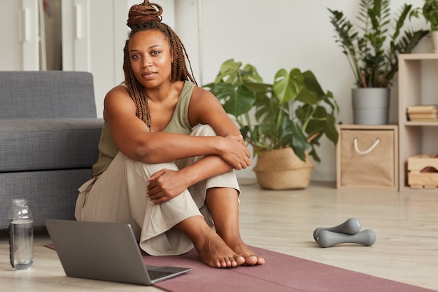 Portret van Afrikaanse vrouw zittend op de vloer met laptop en kijken naar sportoefeningen online voor sporttraining thuis