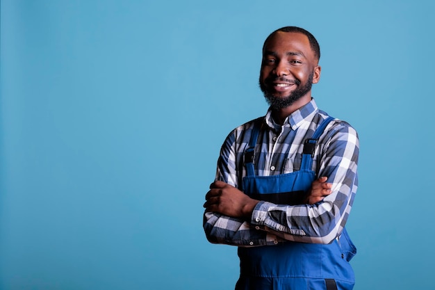 Portret van Afro-Amerikaanse bouwer in uniform poseren vol vertrouwen tegen blauwe achtergrond. Glimlachende bouwprofessional in overall permanent met gekruiste armen in studio.